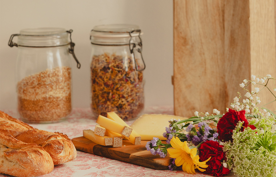 Plateau avec baguettes de pain, morceaux de fromage sur une planche en bois, deux bocaux en verre contenant des céréales et des herbes séchées, et un bouquet de fleurs colorées sur une table à nappe fleurie.