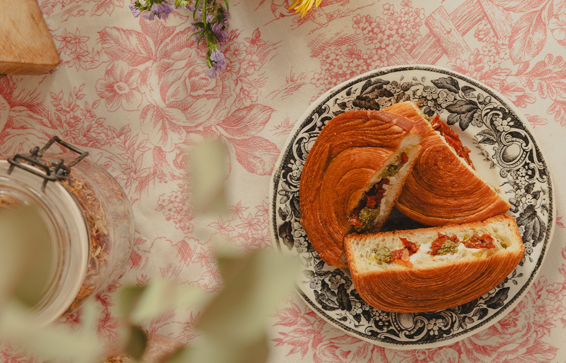Trois sandwichs en croissant avec garniture de fromage, pesto et tomates séchées sur une assiette décorative noire et blanche, posée sur une nappe à motifs floraux rouges.