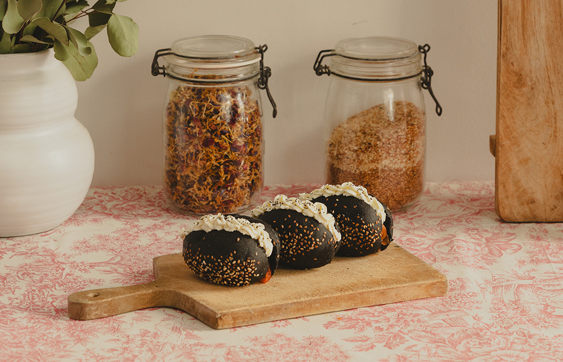 Trois petits pains noirs avec graines de sésame et garniture blanche sur une planche en bois, devant deux pots en verre remplis d'herbes et de graines.