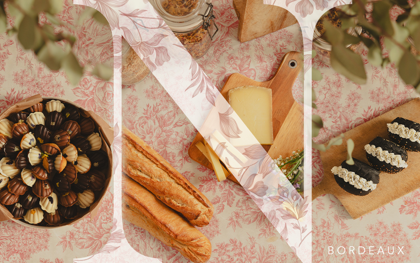 Plateau avec assortiment de chocolats, deux baguettes de pain, fromage sur planche en bois, et trois pâtisseries noires avec crème, sur une nappe fleurie.